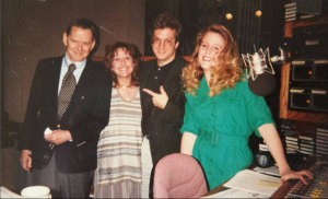 image of the actor Tony Randall standing next to a short woman in her thirties, a man in his 40s with glasses, and a woman in her 20s. They are in a radio station studio with a microphone in the foreground